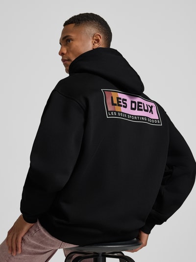 Męski model ma na sobie Hoodie mit Logo-Print Modell 'Chase' od Les Deux z czystej bawełny, luźny krój, czarna kolorystyka, lekko opadające ramiona, nadruk logo, kaptur z troczkiem, kieszeń typu kangurka; zdjęcie w ujęciu z tyłu.