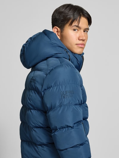 Männermodel trägt Steppjacke mit Label-Stitching und Kapuze von Superdry in Blau, gesteppte Oberfläche, Nahaufnahme Oberkörper. Herren Steppjacke von Superdry, gerader Schnitt, Kapuze mit Kordelzug, Reißverschluss, Label-Stitching, seitliche Eingrifftaschen, elastische Ärmelabschlüsse, Rückenlänge bei Größe M: 67 cm, Schulterbreite bei Größe M: 48 cm, Brustweite bei Größe M: 58 cm, Ärmellänge bei Größe M: 72 cm.