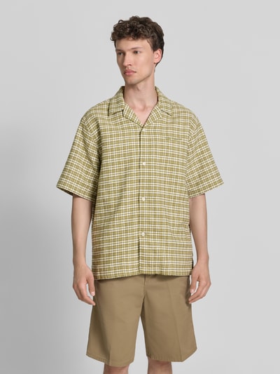 Männermodel trägt Regular Fit Freizeithemd aus reiner Baumwolle von Carhartt Work In Progress mit Strukturmuster, Halbarm, Durchgehende Knopfleiste, Brand-Stitching, Beige- und Grüntöne, Ganzkörperansicht von vorne.