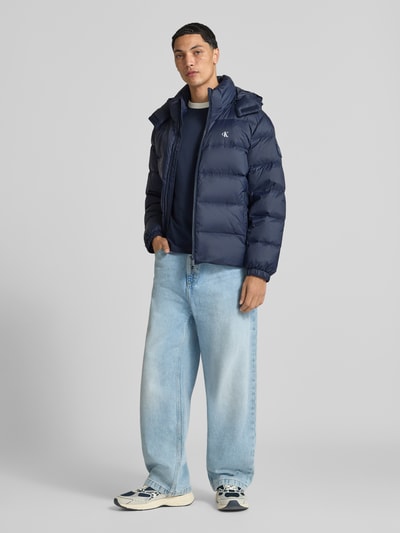 Männermodel trägt Regular Fit Steppjacke mit abnehmbarer Kapuze von Calvin Klein Jeans in Dunkelblau, glatte Textur, Ganzkörperansicht von vorne.