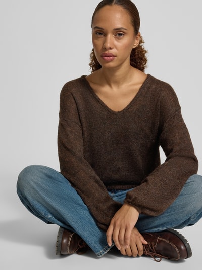 Frauenmodel trägt Regular Fit Pullover mit Woll-Alpaka-Anteil Modell 'Katie' von FREE/QUENT, braun, Strick-Optik, V-Ausschnitt, Rüschenbesatz, Knopfleiste, kombiniert mit blauer Jeans, Ganzkörperansicht von vorne.