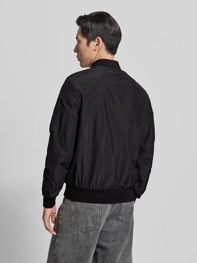 Männermodel trägt Bomberjacke mit Label-Patch und Eingrifftaschen von Original Bombers in Schwarz, glatte Struktur, Ganzkörperansicht Rückansicht, Gerader-Schnitt, Reißverschluss, Reißverschlusstasche am Ärmel, Druckknopfverschluss, Gerippte-Abschlüsse, Rückenlänge bei Größe M 67 cm, Schulterbreite 43 cm, Brustweite 53 cm, Ärmellänge 68 cm.
