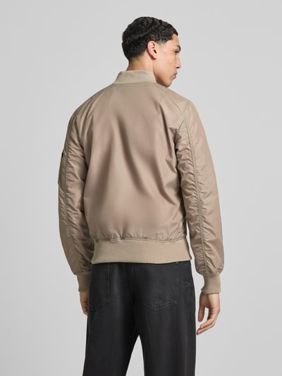 Męski model nosi Regular Fit Jacke mit Stehkragen od Alpha Industries, beżowy kolor, gładka faktura, długość pleców 66 cm, szerokość ramion 48 cm, szerokość klatki piersiowej 54 cm, długość rękawa 68 cm, kieszeń na ramieniu, kieszenie boczne, zamek błyskawiczny, elastyczne wykończenia, widok całej sylwetki od tyłu