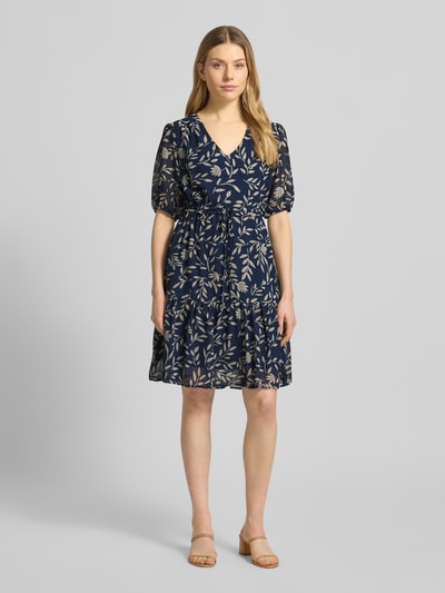 Modelka kobieca nosi Knielanges Kleid mit Kordelzug an der Taille od Apricot, granatowa sukienka z beżowym wzorem liści, fason taliowany, długość do kolan, dekolt w serek, elastyczne zakończenia rękawów, rękaw o dł. 1/2, wiązanie w talii, dół obszyty falbaną, długość tyłu w rozmiarze S: 93 cm, szerokość w rozmiarze S: 43 cm, długość rękawów w rozmiarze S: 30 cm, widok całej sylwetki z przodu.