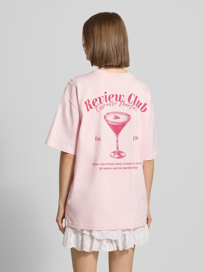 Vrouwenmodel draagt Oversized T-shirt met statementprint van REVIEW in lichtroze, puur katoen, oversized fit, ronde hals, ruglengte bij maat S 70 cm, geheel van achteren gezien.