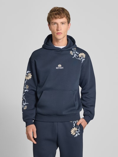 Männermodel trägt Relaxed Fit Hoodie mit Känguru-Tasche von REVIEW in Dunkelblau mit floraler Stickerei, Baumwoll-Mix, Ganzkörperansicht von vorne.