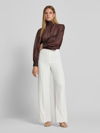 Frauenmodel trägt Regular Fit Bluse aus Viskose-Mix mit Stehkragen von Christian Berg Cocktail, semitransparent, langärmelig, braun, eingefasste Ärmelabschlüsse, kombiniert mit heller Hose, Ganzkörperansicht von vorne.