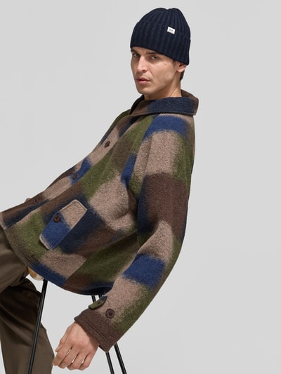 Männermodel trägt Beanie aus Woll-Mix von Forét, Dunkelblau, Ripp-Optik, breiter Umschlag, Label-Patch, Logo-Schriftzug, Nahaufnahme Oberkörper.