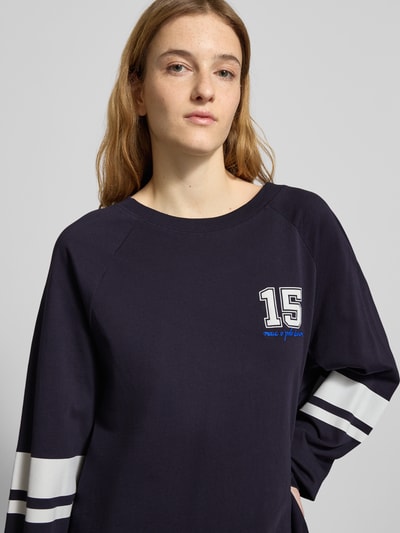 Modelka kobieca nosi Loose Fit Langarmshirt aus reiner Baumwolle od Marc O'Polo Denim, ciemnogranatowy kolor, nadruk z liczbą 15, białe kontrastowe paski na rękawach, materiał bawełniany, widok z przodu, zbliżenie na górną część ciała.