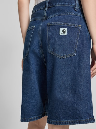 Frauenmodel trägt Relaxed Fit Jeans Shorts aus reiner Baumwolle Modell 'Brandon' von Carhartt Work In Progress, dunkles Blau, Label-Patch, 5-Pocket-Design, Knopf- und Reißverschluss, Damen Jeans Shorts, Ganzkörperansicht Rückansicht.