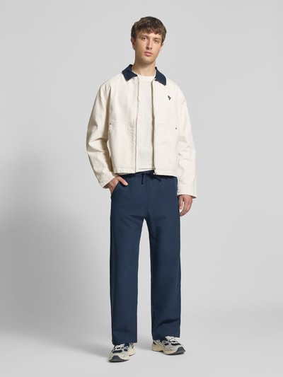 Männermodel trägt Regular Fit tussenjack in denimlook van REVIEW in crème met donkerblauwe platte kraag, puur katoen, ritssluiting, gedeelde kangoeroezak, ruglengte maat M 66 cm, schouderbreedte 50 cm, borstwijdte 59 cm, mouwlengte 66 cm, ganzer lichaamsaanzicht van voren.