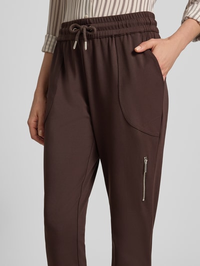 Frauenmodel trägt Jogger Pants mit Reißverschlusstasche Modell 'Jill' von monari in Braun, seitliche Eingrifftaschen, Nahaufnahme Oberkörper, Material-Mix mit Stretch-Anteil, elastischer Bund mit Kordelzug, elastischer Saum, angedeutete Gesäßleistentaschen.