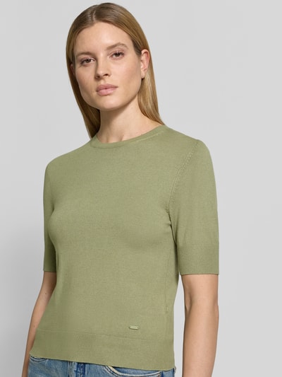 Modelka kobieca ma na sobie Slim Fit T-Shirt aus Viskose-Mix od s.Oliver RED LABEL w kolorze oliwkowym, imitacja dzianiny, aplikacja z metką, okrągły dekolt, krótki rękaw, zakończenia prążkowane, wykończenie drobny splot, mieszanka wiskozy, zbliżenie tułowia z przodu.
