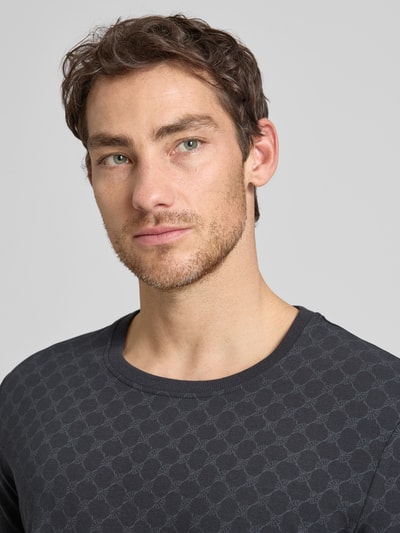 Männermodel trägt T-Shirt mit Logo-Muster und Rundhalsausschnitt von JOOP! Collection aus Baumwoll-Elasthan-Mix, Nahaufnahme Oberkörper. Regular-Fit, Rückenlänge bei Größe M: 68 cm, dunkle Farbe mit strukturierter Textur.