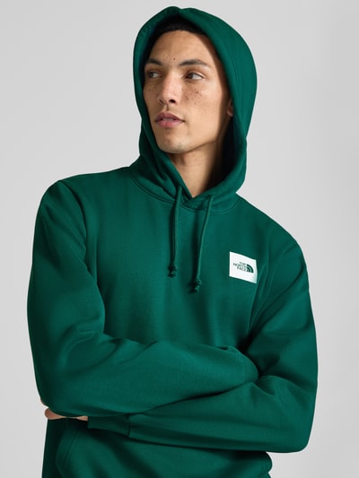 Männermodel trägt Regular Fit Hoodie mit Känguru-Tasche von The North Face in Grün mit Kapuze und Logo, Nahaufnahme Oberkörper. Herren Hoodie, gerippte Abschlüsse, Rückenlänge 67 cm, Ärmellänge 68 cm.