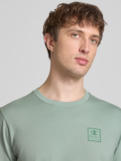 Männermodel trägt T-Shirt mit Logo und Rundhalsausschnitt von CHAMPION aus Reine Baumwolle, Regular-Fit, Rückenlänge 68 cm, Nahaufnahme Oberkörper.