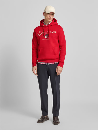 Męski model nosi czerwony Hoodie mit Label-Stitching und Känguru-Tasche od Gant z bawełnianego miksu, regular fit, zdjęcie całej sylwetki z przodu.
