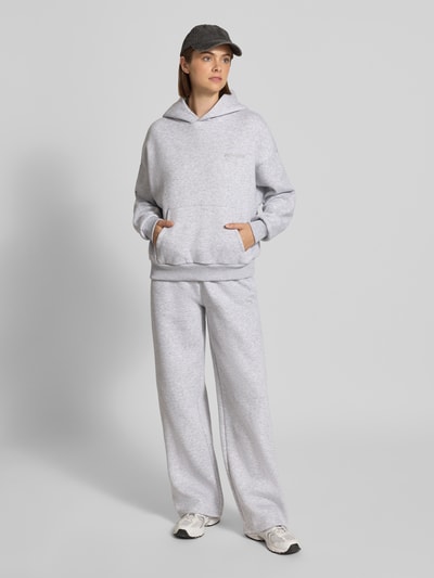 Kobiece model prezentuje Relaxed Fit Basic Hoodie in grau melange od REVIEW, mieszanka bawełny, krój relaxed fit, kaptur, nadruk z logo, kieszeń kangurka, prążkowane wykończenia, szary melanż, widok całej sylwetki z przodu.