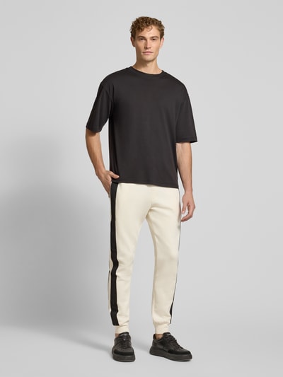Männermodel trägt Sweatpants mit Bordüre von Armani Exchange in Creme mit schwarzer Seitenlinie, gerader Schnitt, Ganzkörperansicht von vorne. Elastischer Baumwoll-Mix, Label-Print an der Bordüre, Elastikbund mit Tunnelzug, seitliche Eingrifftaschen, Gesäßtasche, Innenbeinlänge bei Größe M: 79 cm.