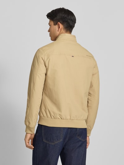 Męski model nosi kurtkę 'Jacke mit Stehkragen und Eingrifftaschen' od Superdry, kolor beżowy, krój regular fit, zamek błyskawiczny, wpuszczane kieszenie, długość tyłu w rozmiarze M: 70 cm, szerokość ramion w rozmiarze M: 44 cm, szerokość klatki piersiowej w rozmiarze M: 41 cm, długość rękawów w rozmiarze M: 68 cm, gładka faktura, widok całej sylwetki z tyłu.