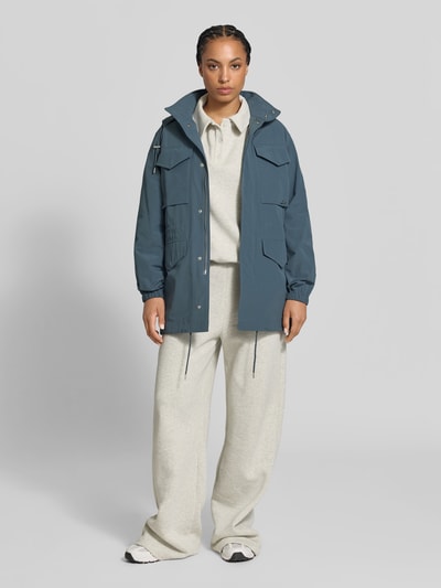 Frauenmodel trägt Windbreaker mit Pattentaschen von Kenzo in Blau, lockere Passform, abnehmbare Kapuze, beige Hose, Ganzkörperansicht von vorne. Rückenlänge 74 cm, Brustweite 50 cm, Ärmellänge 65 cm, glänzende glatte Textur, Reißverschluss mit Abdeckleiste, Taille und Bund mit Tunnelzug, Pattentaschen.