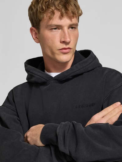 Männermodel trägt Oversized Hoodie aus Baumwoll-Mix von PEGADOR in Schwarz mit Logo-Stickerei, Nahaufnahme Oberkörper.