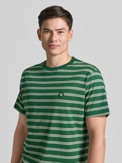 Męski model nosi Relaxed Fit T-Shirt mit Logo-Stitching Modell 'Easton' od Wood Wood, bawełna, zielone w białe paski, krój relaxed fit, widok z przodu, zbliżenie na górną część sylwetki.