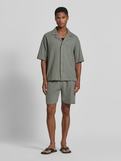 Męski model nosi Relaxed Fit Freizeithemd mit Strukturmuster von Drykorn z bawełny, kołnierz z połami, krótki rękaw, listwa guzikowa, szara faktura, całkowity widok z przodu. Długość tyłu w rozmiarze M: 69 cm, szerokość w rozmiarze M: 60 cm, długość rękawów w rozmiarze M: 26 cm.