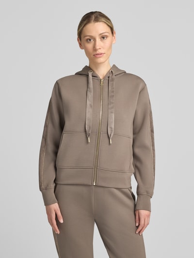 Frauenmodel trägt Relaxed Fit Sweatjacke mit Viskose-Anteil von GUESS in Taupe, Reißverschluss, Kapuze, Kängurutasche, Ganzkörperansicht von vorne.