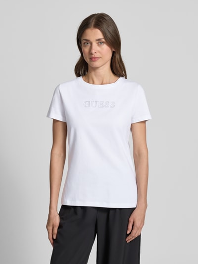 Frauenmodel trägt T-Shirt mit Logo Modell 'BRIANA' von Guess Activewear, weiß, Rundhalsausschnitt, Ziersteinbesatz, Ganzkörperansicht von vorne.
