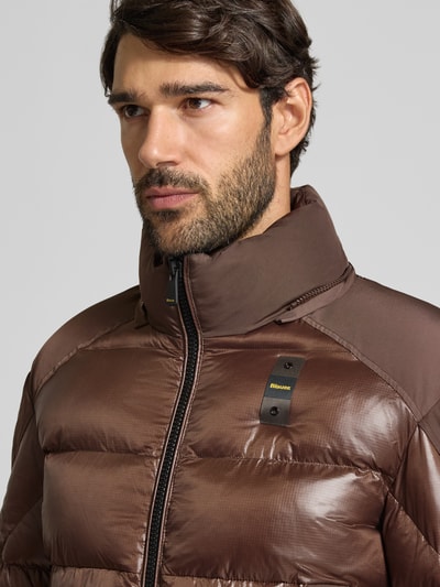 Mężczyzna nosi Steppjacke mit Zweiwege-Reißverschluss Modell 'KIERNAN' od Blauer USA, brązowy kolor, gładka powierzchnia, metka z logo, krój regular fit, kurtka z kapturem, widok na górną część tułowia, zbliżenie. Szerokość w rozmiarze M: 59 cm, długość tyłu: 67 cm, szerokość ramion: 49 cm, długość rękawa: 65 cm.