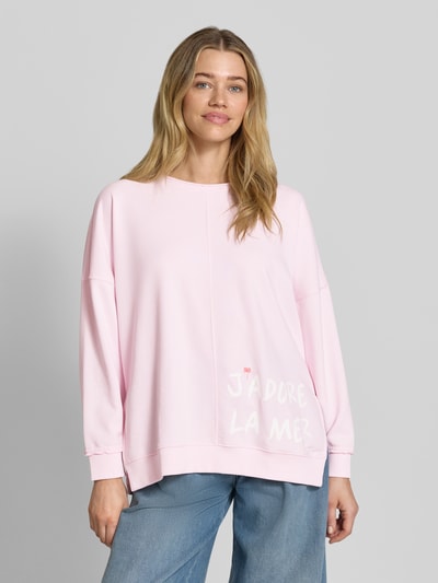 Frauenmodel trägt Oversized Sweatshirt mit Statement-Print und Rollsaum von Smith and Soul aus Baumwoll-Mix mit Modal-Anteil in Hellrosa, Rundhalsausschnitt, überschnittene Schultern, weiße Schrift, glatte Struktur, Ganzkörperansicht von vorne.