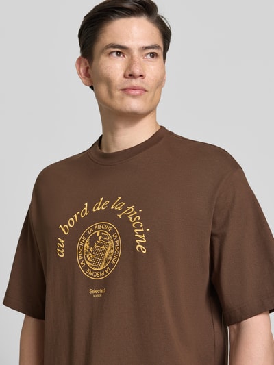 Männermodel trägt Relaxed Fit T-Shirt aus reiner Baumwolle Modell 'BOXYNORMAN GRAPHIC' von SELECTED HOMME in Braun mit gelbem Motiv-Print, Rundhalsausschnitt und kurzen Ärmeln, Jersey-Textur, Nahaufnahme Oberkörper.
