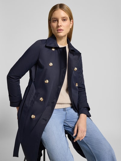 Frauenmodel trägt dunkelblauen Trenchcoat mit Ärmelriegeln von Christian Berg Woman Selection, beiger Pullover, blaue Jeans, Nahaufnahme Oberkörper. Trenchcoat in taillierter Passform, doppelreihiger Knopfleiste, Taillengürtel mit Dornschließe, glänzende Knöpfe, glatte Textur.