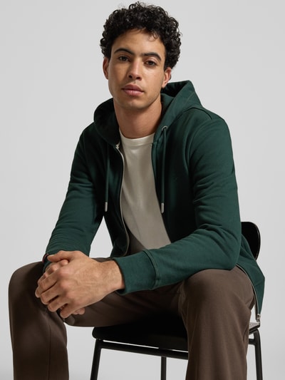 Männermodel trägt Regular Fit Sweatjacke mit Logo-Stitching von Superdry aus grünem Baumwoll-Mix, Nahaufnahme Oberkörper.
