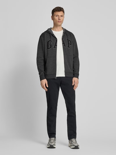 Männermodel trägt Sweatjacke mit Logo und Känguru-Tasche von GAP aus Baumwoll-Mix, Regular-Fit, dunkel, Ganzkörperansicht von vorne.