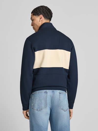 Męski model nosi bluzę rozpinaną z mieszanki bawełny z dodatkiem streczu Helly Hansen, prosty krój, model 'KOSTER', stójka, zamek błyskawiczny, naszywka z logo, wpuszczane kieszenie, długość tyłu w rozmiarze M: 67 cm, długość rękawów w rozmiarze M: 66 cm, granatowy z beżowym panelem, tekstura gładka, widok całej sylwetki z tyłu.