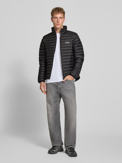 Männermodel trägt Steppjacke mit Label-Print von Helly Hansen in Schwarz mit durchgehendem Reißverschluss, Ganzkörperansicht von vorne. Gerade Passform, Stehkragen, elastische Ärmelabschlüsse, Rückenlänge 72 cm, Schulterbreite 42 cm, Brustweite 54 cm, Ärmellänge 72 cm.