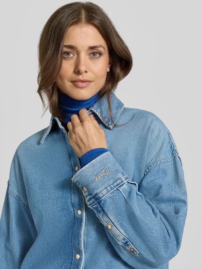 Kobiece model prezentuje Oversized Hemdjacke aus Baumwoll-Denim Modell 'C_OVERSHIRT 4.0' von BOSS Orange, jednokolorowy denim, krój oversized, wykładany kołnierz, listwa z zatrzaskami, długi rękaw, wpuszczane kieszenie, wykończenie: denim, materiał: bawełna, zbliżenie na górną część ciała.