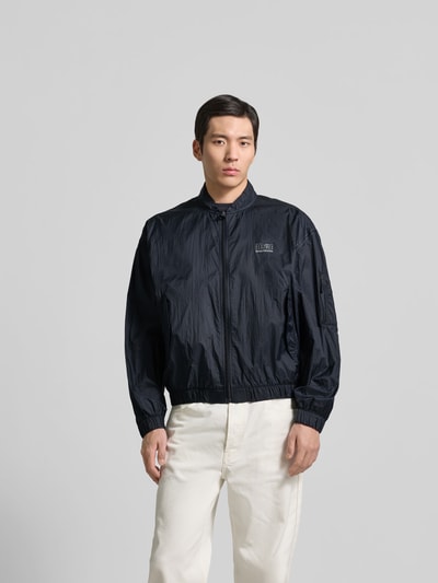 Männermodel trägt Relaxed Fit Jacke mit Logo-Print von MM6 Maison Margiela in Schwarz, Stehkragen, Reißverschluss, elastische Abschlüsse, Ganzkörperansicht von vorne.
