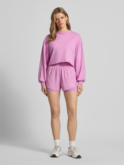 Frauenmodel trägt Shorts im 2-in-1-Look mit Logo von NIKE TRAINING in Rosa, Material-Mix mit Stretch-Anteil, elastischer Bund, Ganzkörperansicht von vorne.