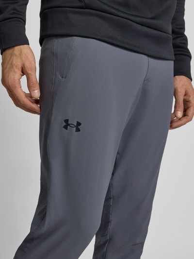 Męski model nosi szare spodnie treningowe z kieszeniami na zamek Traniningshose mit Reißverschlusstaschen od Under Armour, widok z przodu, zbliżenie na nogawki. Regularny krój, elastyczny pas, długość nogawki M: 84 cm.