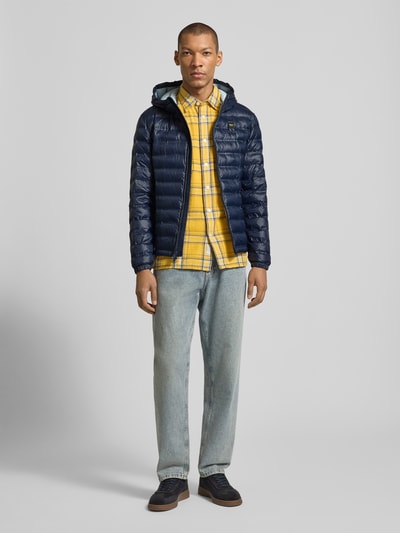 Männermodel trägt Steppjacke mit Zweiwege-Reißverschluss Modell 'Charles' von Blauer Usa in Dunkelblau, Kapuze, Regular-Fit, Label-Detail, kombiniert mit gelb kariertem Hemd und Jeans, Rückenlänge 65 cm, Schulterbreite 47 cm, Brustweite 55 cm, Ärmellänge 65 cm, Ganzkörperansicht von vorne.