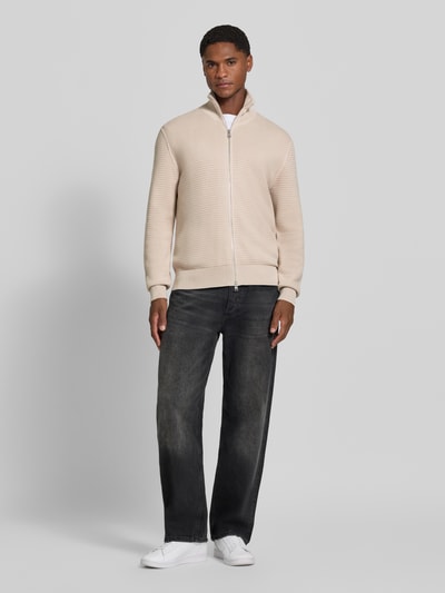 Mężczyznęmodel nosi Regular Fit Strickjacke aus reiner Baumwolle von Marc O'Polo w kolorze beżowym, czarne jeansy, białe buty, sweter z dzianiny, Ganzkörperansicht von vorne, sweter z bawełny, Herren Pullover von Marc O'Polo.