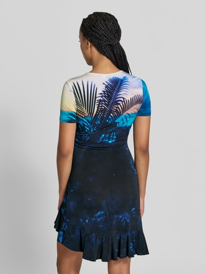 Frauenmodel trägt T-Shirt-Kleid aus Viskose-Mix von Rabanne mit Allover-Foto-Print in Blau- und Naturtönen, asymmetrischer Saum mit Rüschen, eng anliegende Passform, kurzärmlig, Ganzkörperansicht Rückansicht.