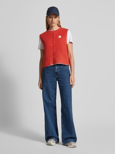 Frauenmodel trägt Regular Fit Weste mit Alpaka-Woll-Anteil von American Vintage, Strick-Optik, gerippte Abschlüsse, Rundhalsausschnitt, Knopfleiste, Label-Detail, Material-Mix, Rückenlänge bei Größe S 49 cm, kräftiges Rot, kombiniert mit weißem T-Shirt und blauer Hose, Ganzkörperansicht von vorne.