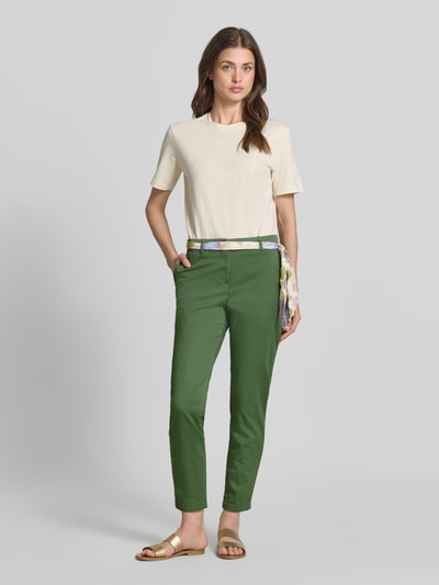 Kobieta nosi Slim Fit Chino mit Viskose-Anteil von Christian Berg Woman z mieszanki bawełny i wiskozy, krój slim fit, skrócony, z odpinaną materiałową tasiemką, zapięcie na guzik i zamek błyskawiczny, zielony kolor, gładka faktura, pełny widok z przodu.