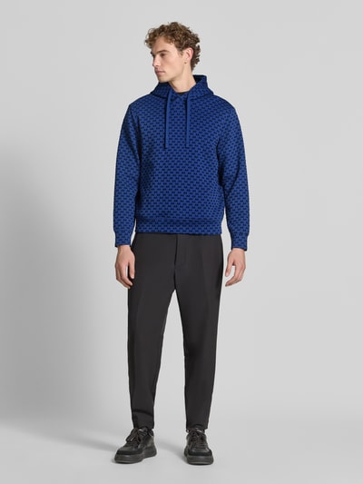 Männermodel trägt Regular Fit Hoodie aus Baumwoll-Mix von Armani Exchange mit Allover-Logo-Print in Blau und Schwarz, Ganzkörperansicht von vorne.