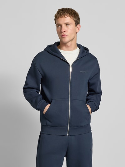 Männermodel trägt Regular Fit Sweatjacke mit Reißverschluss von Review aus dunkelblauem Baumwoll-Mix mit Logo-Print, Kapuze, Kängurutasche, elastischen Abschlüssen, Ganzkörperansicht von vorne.