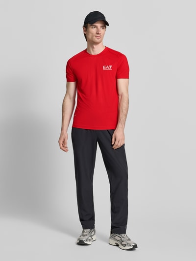 Männermodel trägt T-Shirt mit Label-Print von EA7 Emporio Armani in Rot, Material-Mix mit Stretch, Slim Fit, Ganzkörperansicht von vorne.